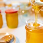 An unknown person pours sweet golden honey into transparent empty clean jars standing on a wooden white large table on a bright sunny day